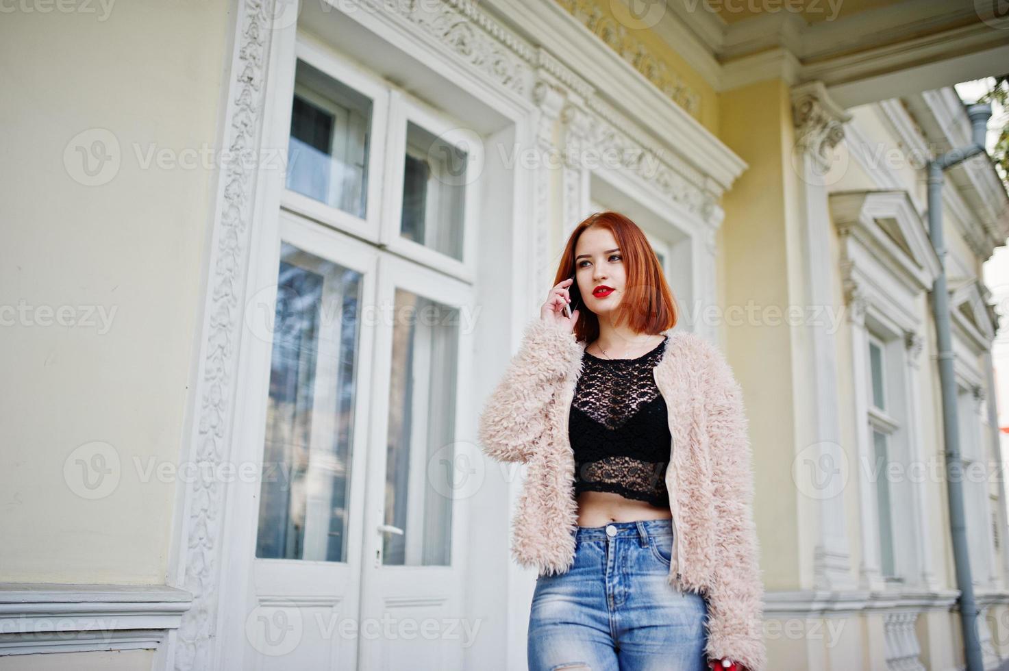 chica pelirroja con bolso rojo posado cerca de casa vintage y hablando por teléfono móvil. foto