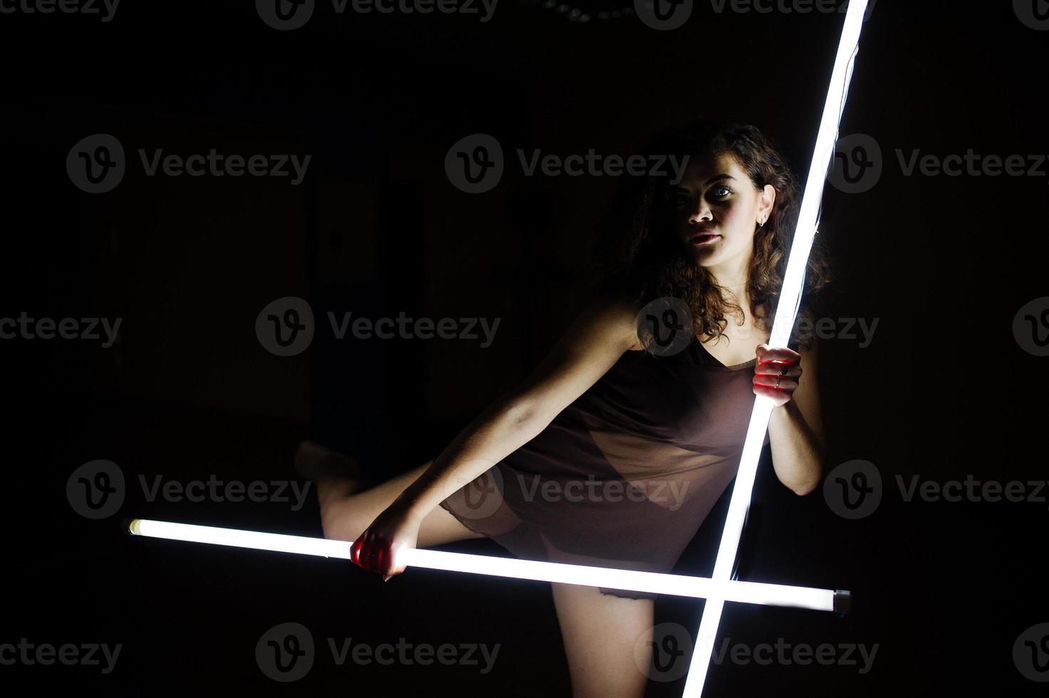 Curly brunette plus size model with two long led lamp at black room. photo