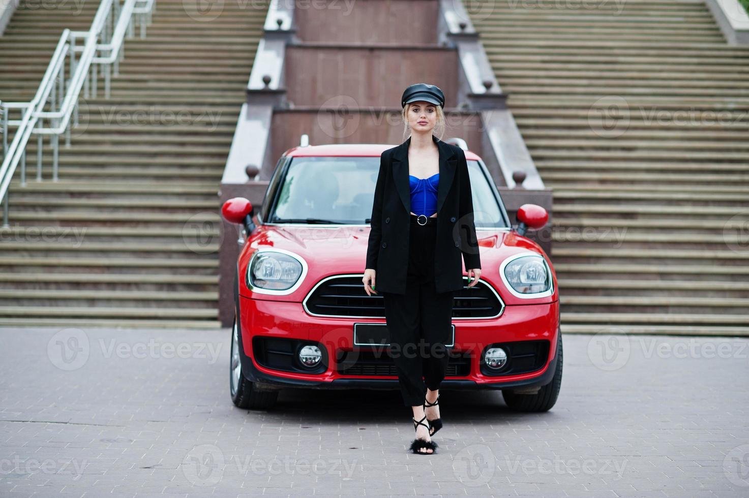 retrato de una hermosa modelo rubia de moda sexy con gorra y corsé negro y azul, con maquillaje brillante cerca del coche rojo de la ciudad. foto