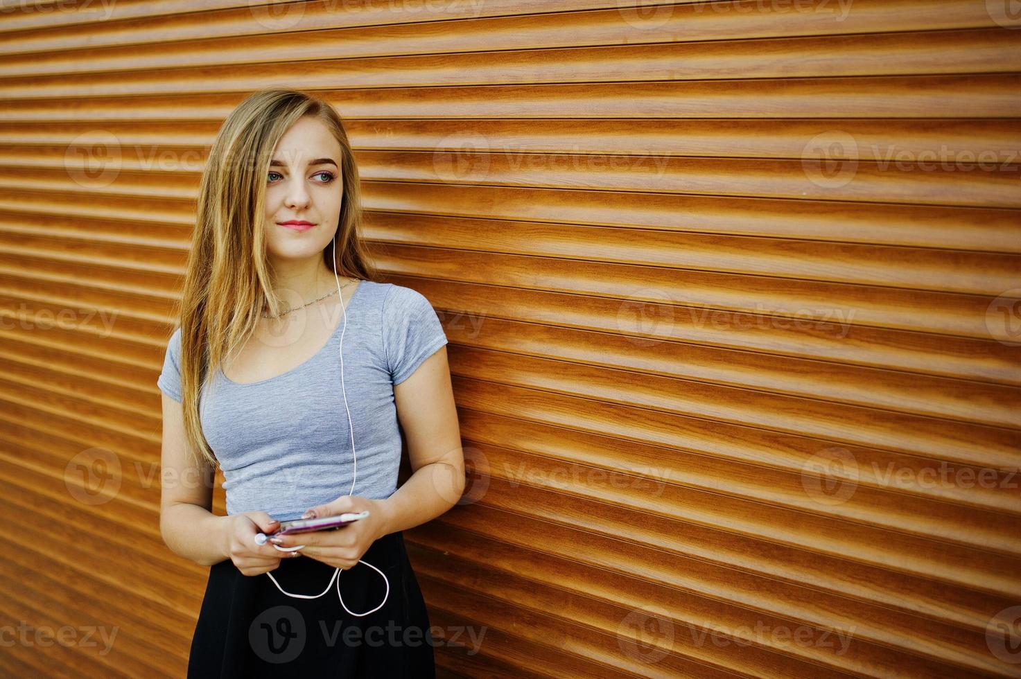 jovencita rubia con falda negra escuchando música desde auriculares de teléfono móvil contra persianas naranjas. foto