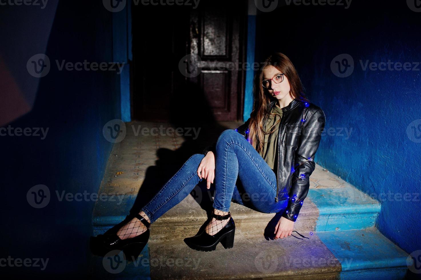 retrato nocturno de modelo de niña con gafas, jeans y chaqueta de cuero, con guirnalda azul sobre ella. foto