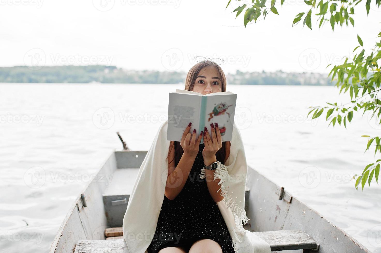 Portrait of an attractive woman wearing black polka dots dress, white shawl and glasses reading a book in a boat on a lake. photo