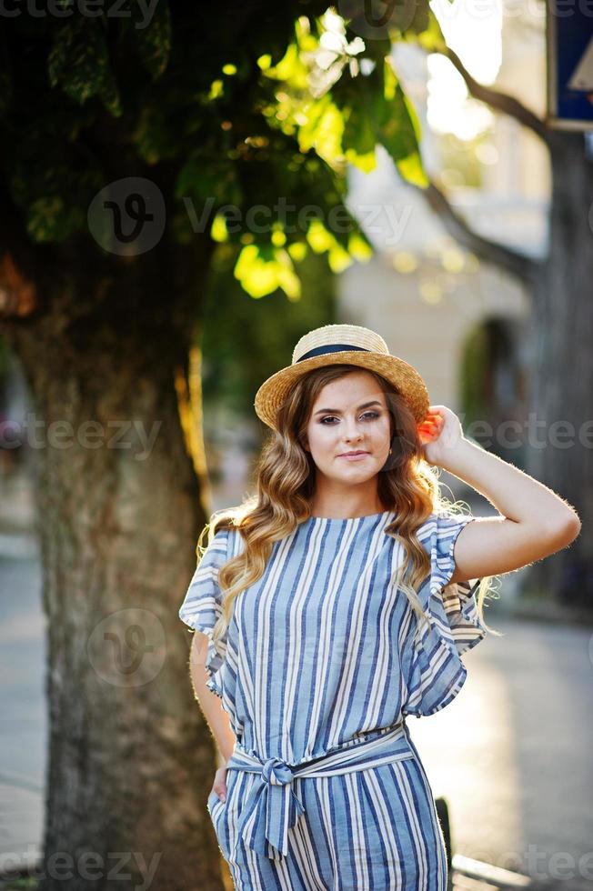 Portrait of a very attractive young woman in striped overall posing with her hat on a pavement in a town with trees in a background. photo