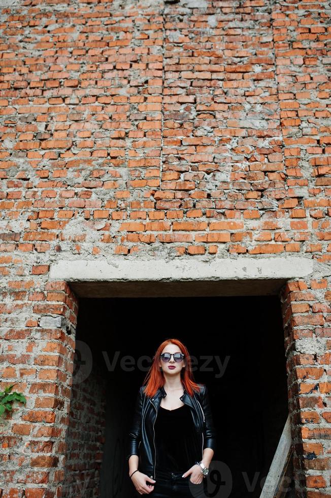 Red haired stylish girl in sunglasses wear in black, against abadoned place with brick walls. photo