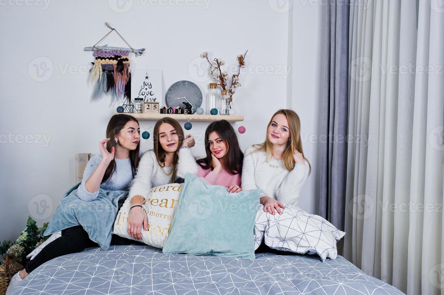 Four cute friends girls wear on warm sweaters and black pants on bed at new year decorated room on studio, play with pillows. photo
