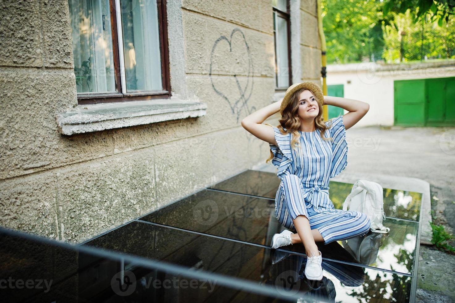 Portrait of a fabulous young woman wearing striped overall and hat sitting on black shiny surface with backpack next to the building. photo