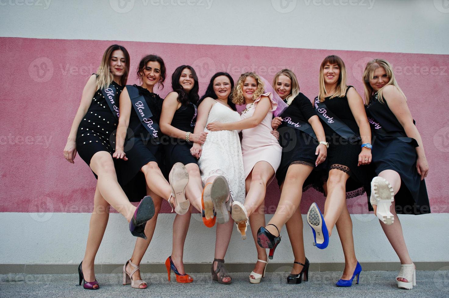 grupo de 8 chicas vestidas de negro y 2 novias en una despedida de soltera posadas contra una pared rosa. foto