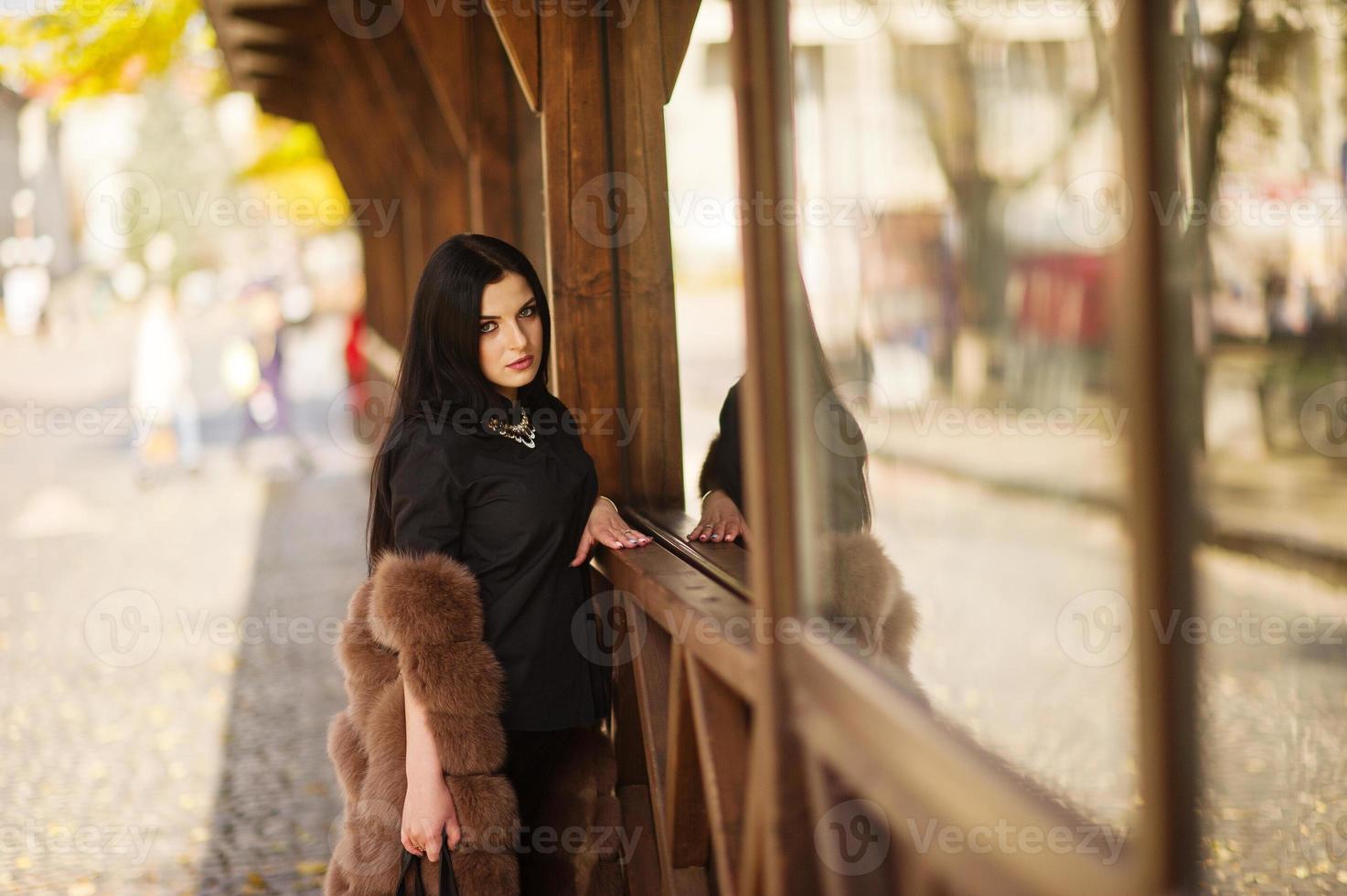 Fashion outdoor photo of gorgeous sensual woman with dark hair in elegant clothes and luxurious fur coat against wooden terrace at autumn city.