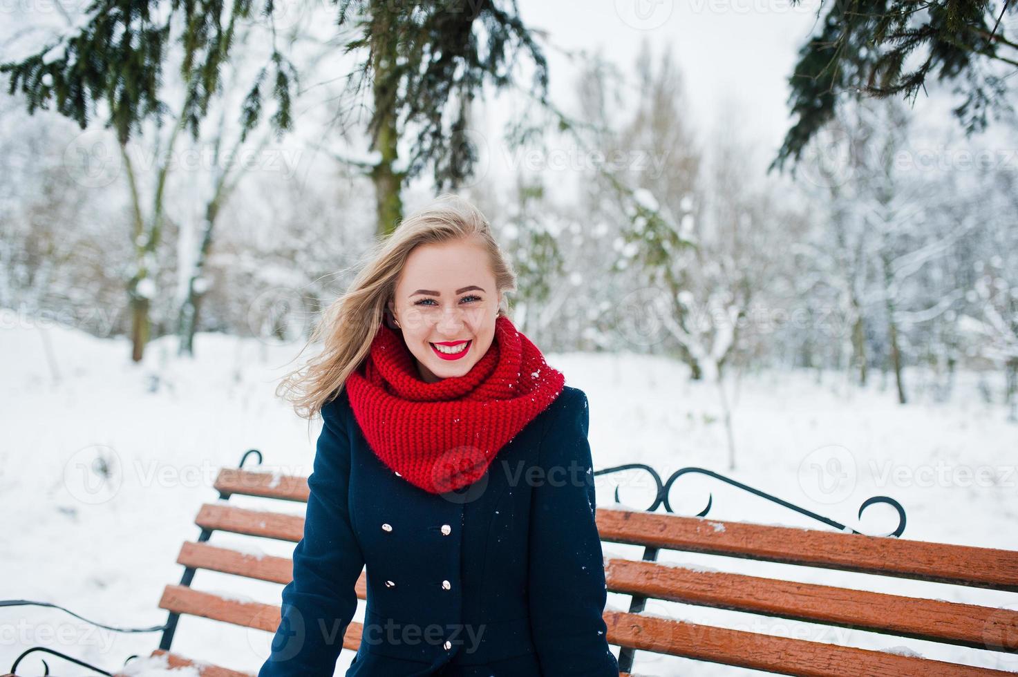 Blonde girl in red scarf and coat sitting at bench on winter day. photo