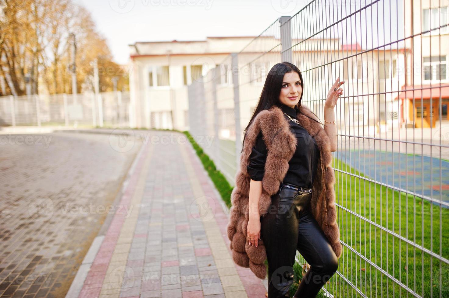 foto de moda al aire libre de una hermosa mujer sensual con cabello oscuro en ropa elegante y lujoso abrigo de piel sin mangas contra la cerca de hierro en la ciudad de otoño.