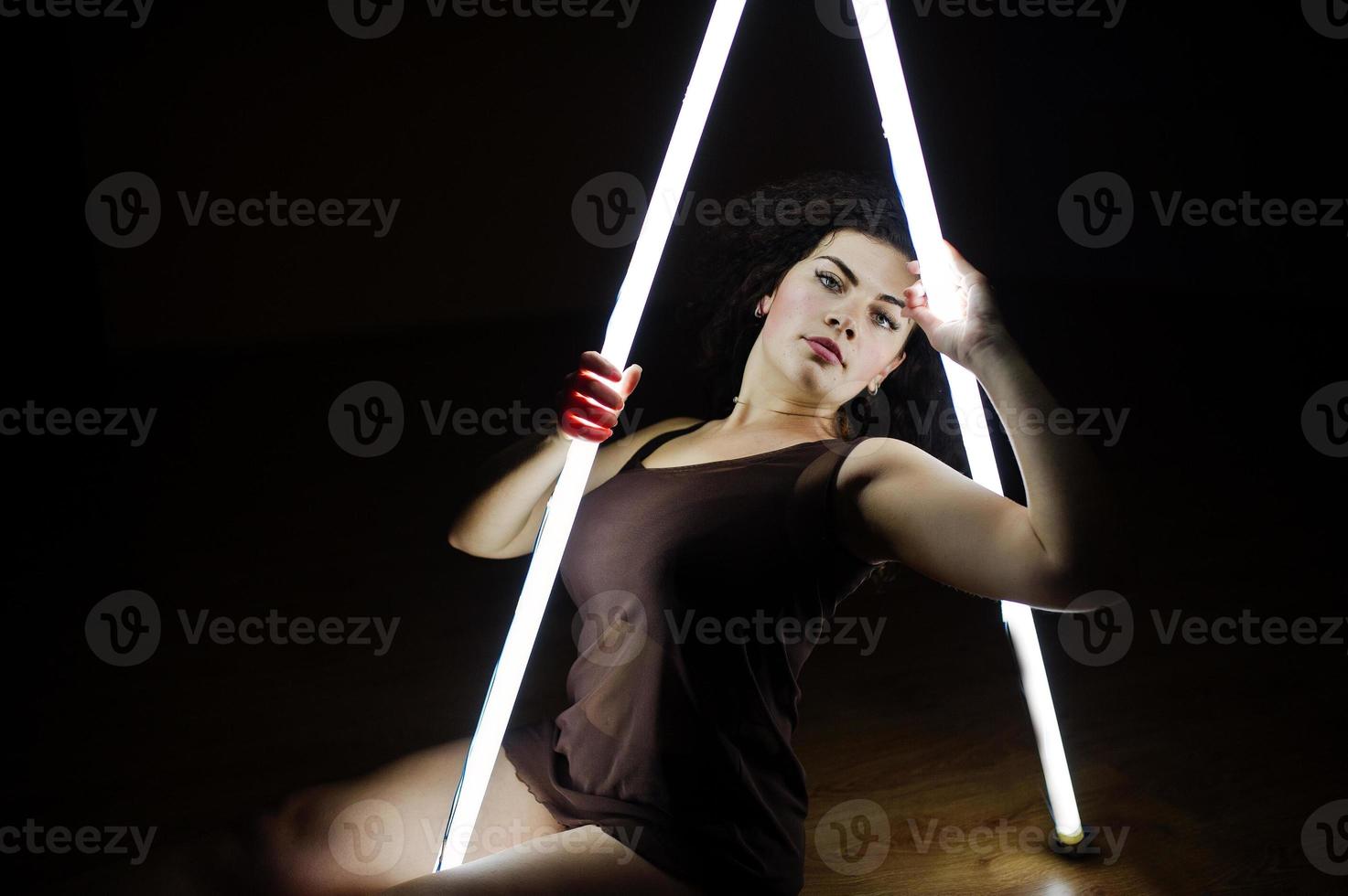 Curly brunette plus size model with two long led lamp at black room. photo