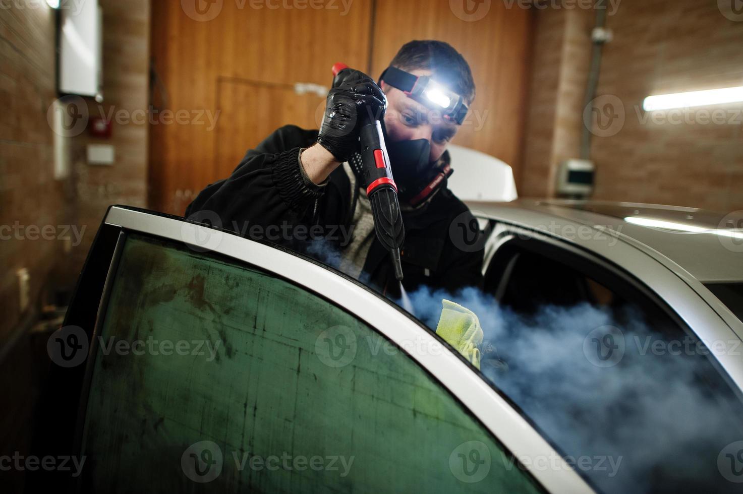 Man in uniform and respirator, worker of car wash center, cleaning car interior with hot steam cleaner. Car detailing concept. photo