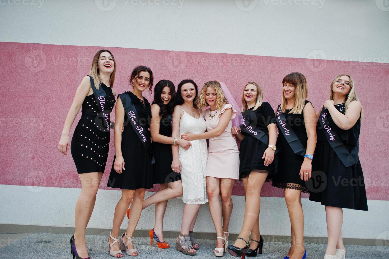 grupo de 8 chicas vestidas de negro y 2 novias en una despedida de soltera posadas contra una pared rosa. foto