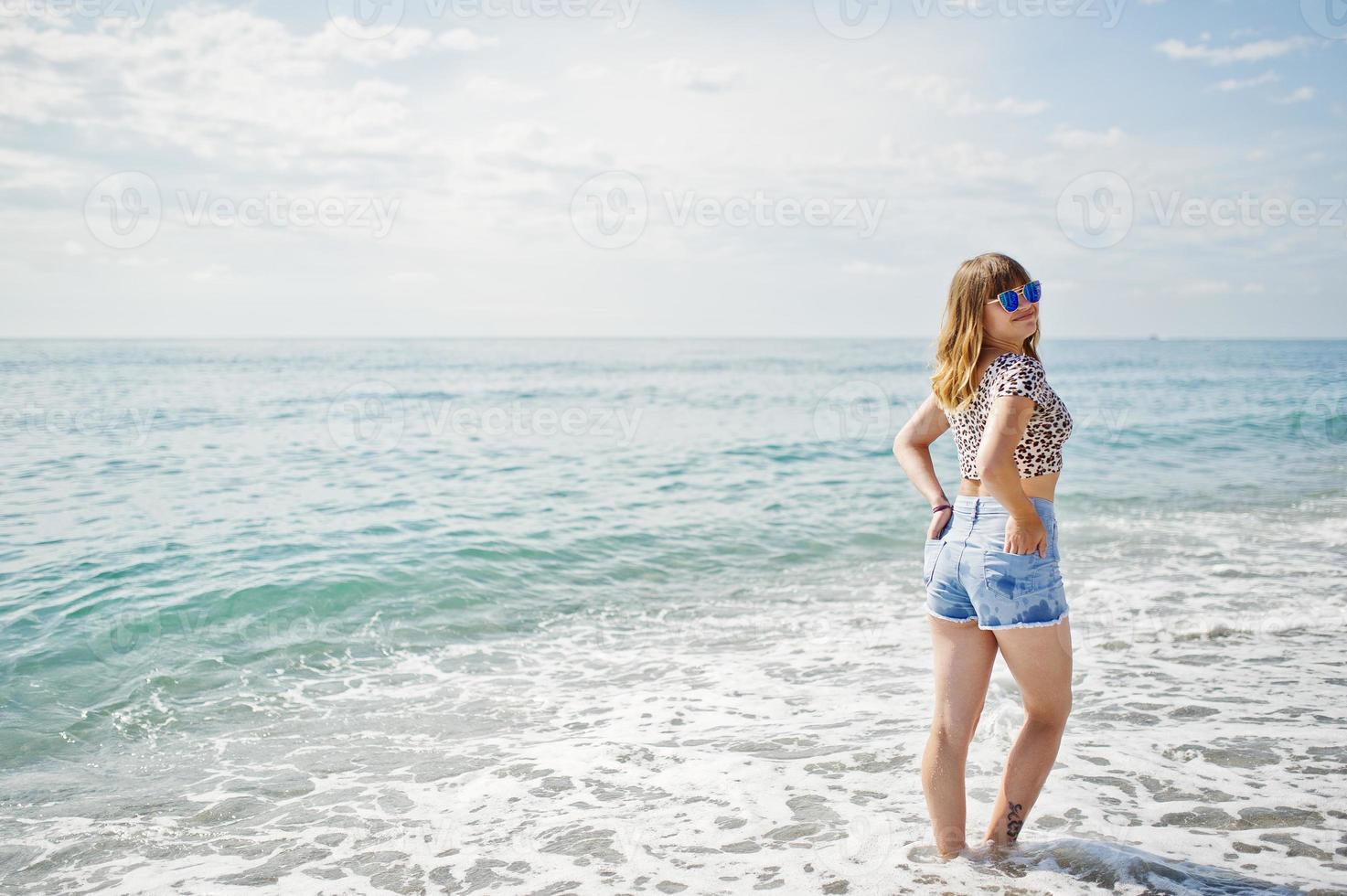 hermosa modelo relajándose en una playa de mar, usando jeans cortos, camisa de leopardo y gafas de sol. foto