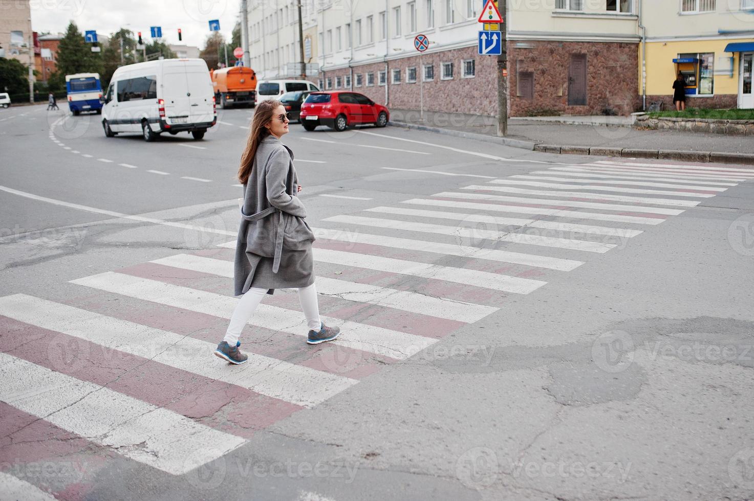 chica de abrigo gris con gafas de sol y bolso caminando por el paso de peatones. foto