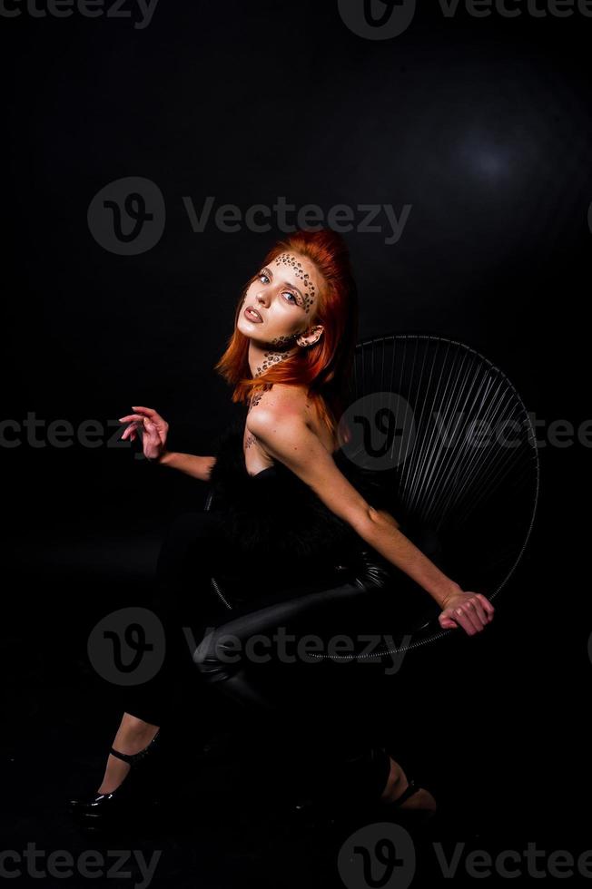 Fashion model red haired girl with originally make up like leopard predator isolated on black. Studio portrait on chair. photo