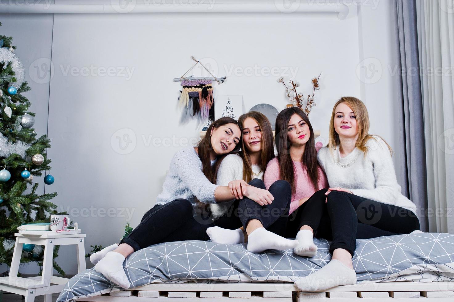 Four cute friends girls wear on warm sweaters and black pants on bed at new year decorated room on studio. photo