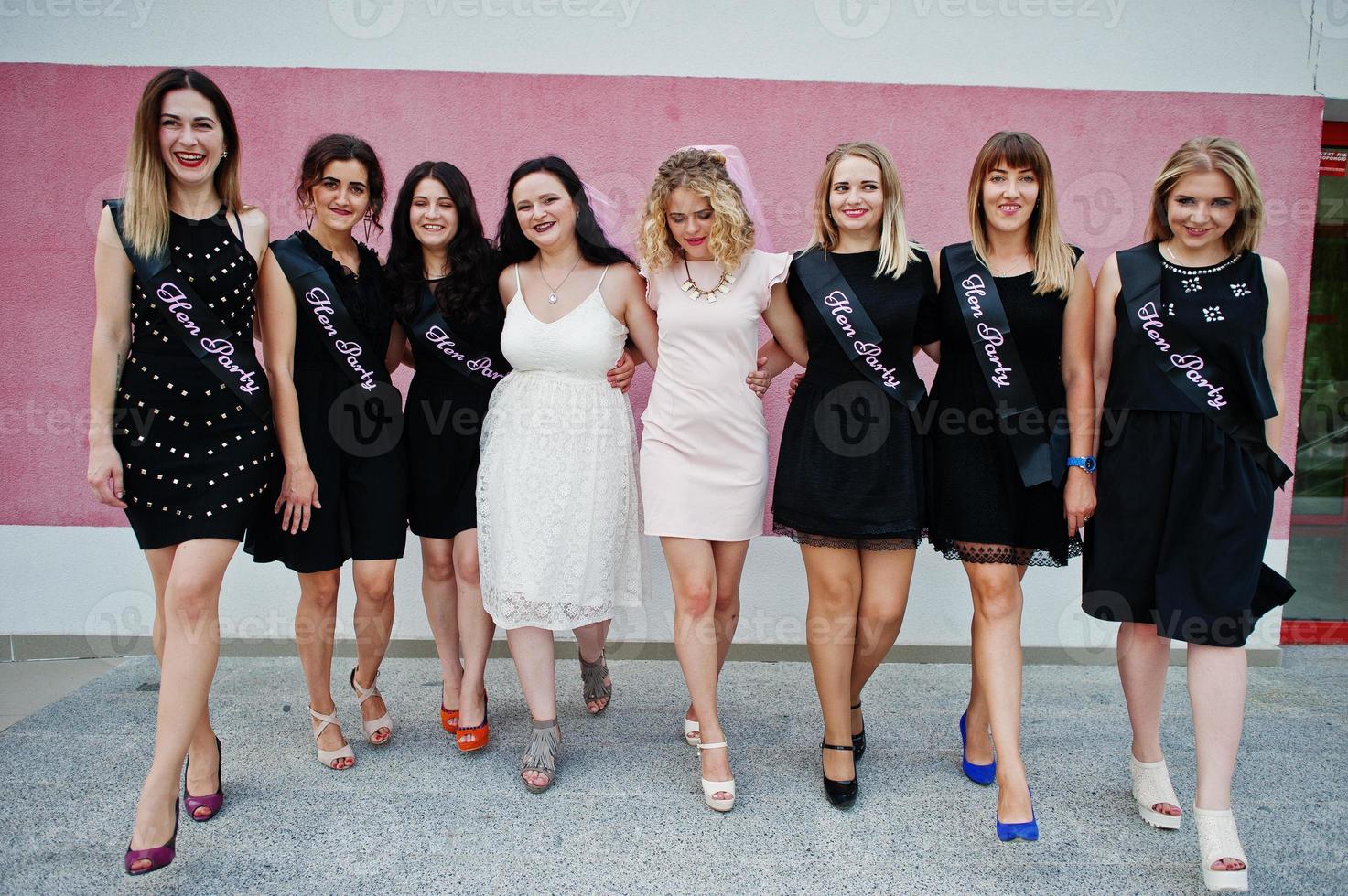 Group of 8 girls wear on black and 2 brides at hen party posed against pink wall. photo