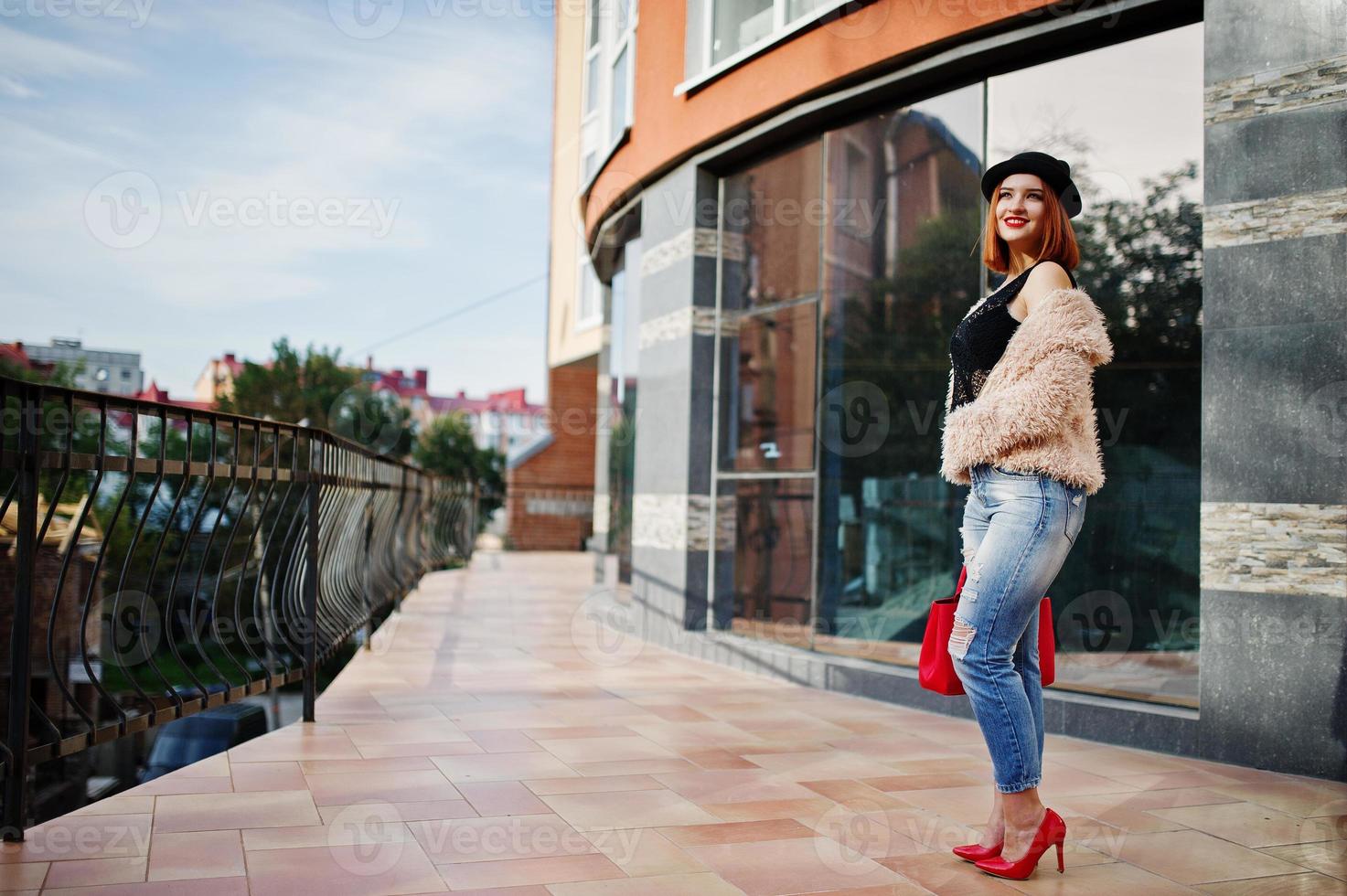 Red haired girl in  hat with red handbag posed near modern house. photo