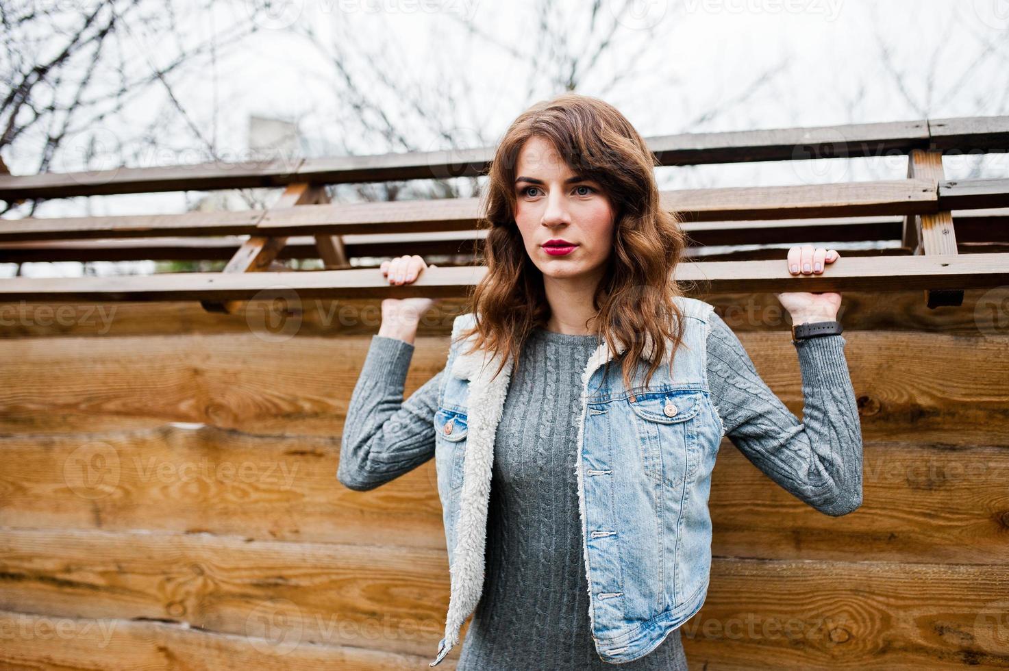 Portrait of brunette curly girl in jeans jacket against wooden wall. photo