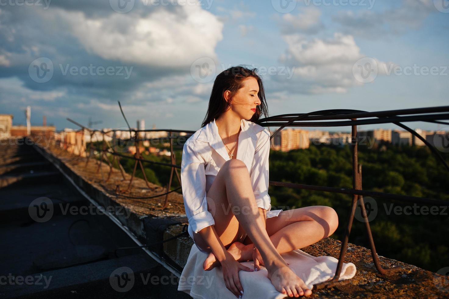 Portrait of a fabulous young woman in white male shirt sitting on the edge of the building roof with a fantastic view of a city in a background. photo