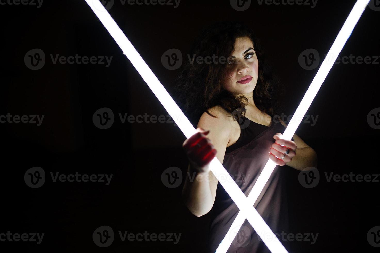 Curly brunette plus size model with two long led lamp at black room. photo