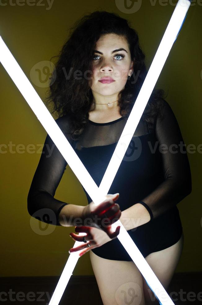 Curly brunette plus size model with two long led lamp against yellow wall. photo