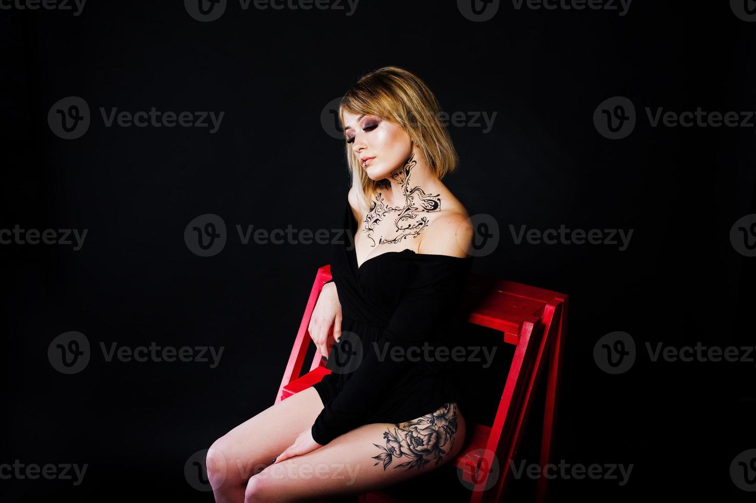 Studio portrait of blonde girl with originally make up on neck and tattoo on thigh, wear on black dress at dark background, sitting on red ladder. photo