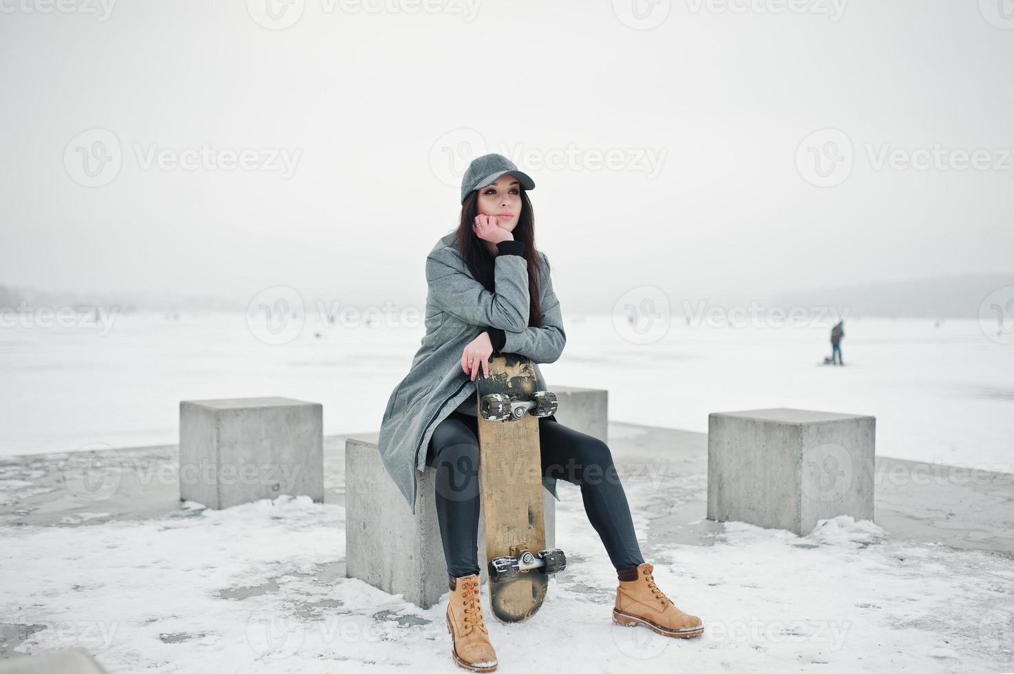 Stylish brunette girl in gray cap, casual street style with skate board on winter day. photo