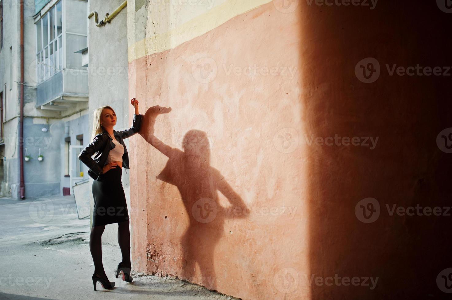 Elegant blonde girl wear on black leather jacket posing at streets of town background old wall with shadows. photo