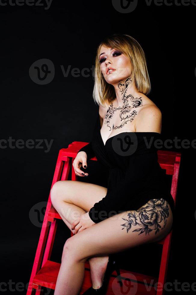 Studio portrait of blonde girl with originally make up on neck and tattoo on thigh, wear on black dress at dark background, sitting on red ladder. photo