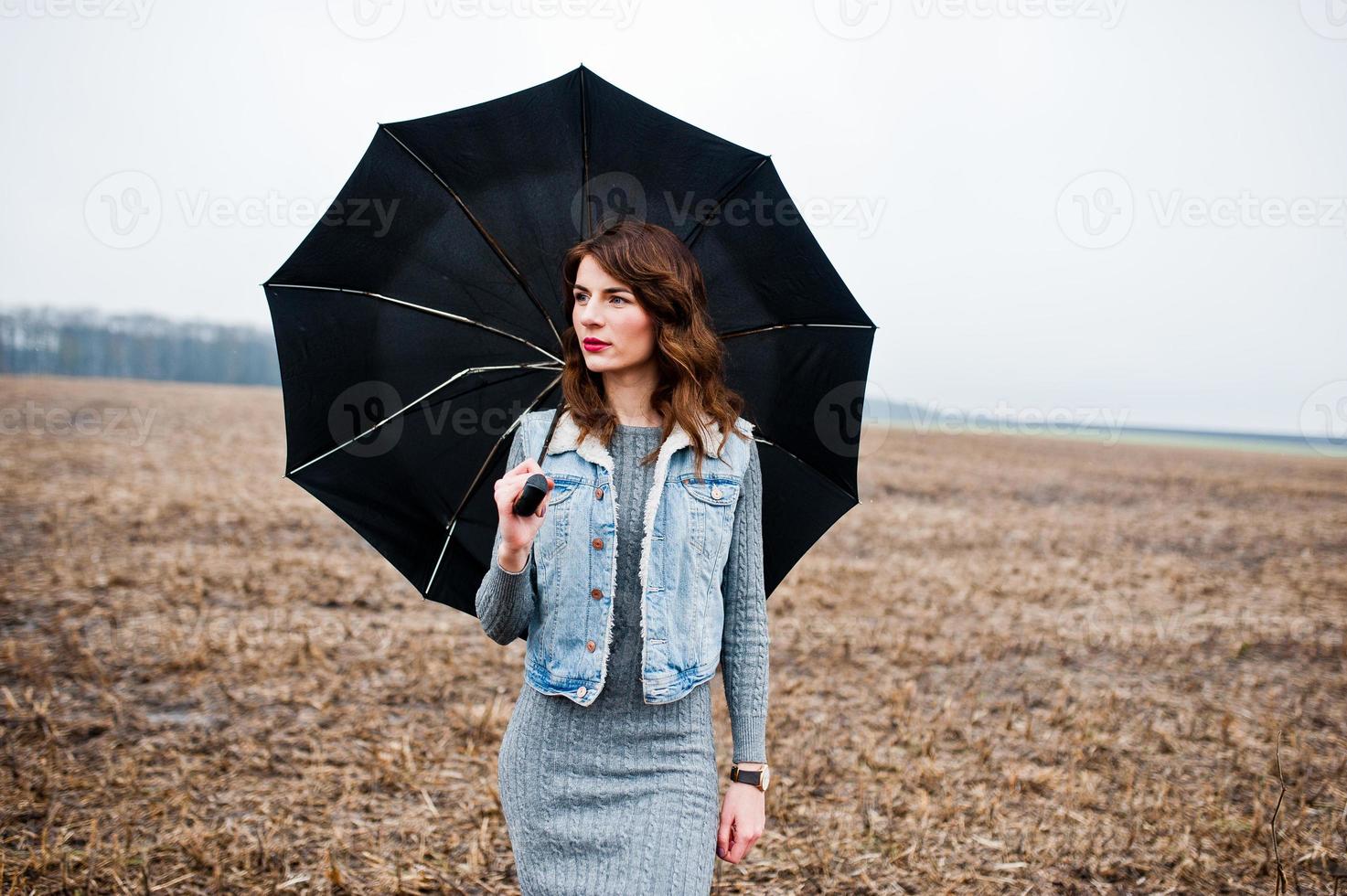 Portrait of brunette curly girl in jeans jacket with black umbrella at field. photo