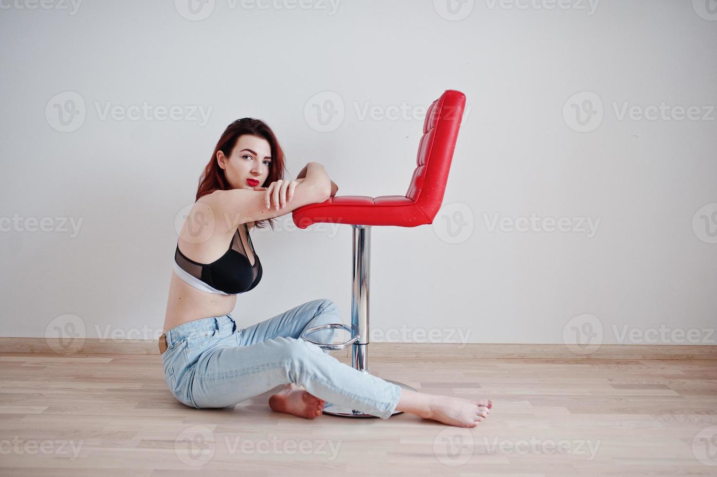 chica pelirroja con un gran busto en sujetador negro y jeans sentada cerca de una silla roja contra una pared blanca en una habitación vacía. foto