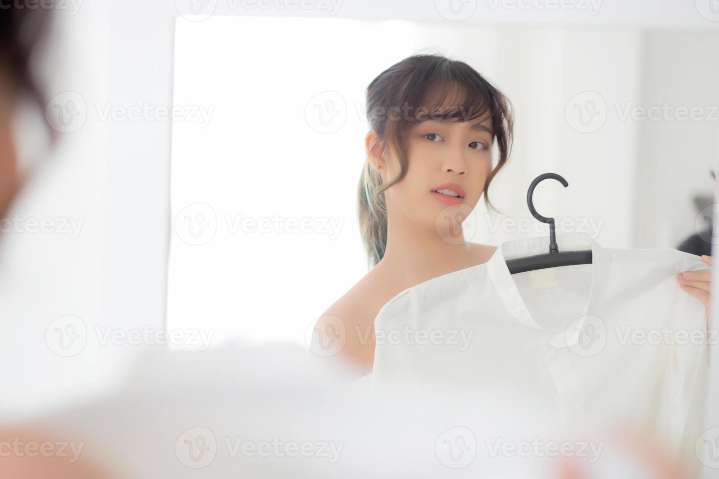 joven mujer asiática sonriendo con ropa tratando de vestirse con un espejo de reflexión moderno y de aspecto en la habitación, la chica elige ropa de moda con un concepto de estilo de vida feliz y alegre. foto