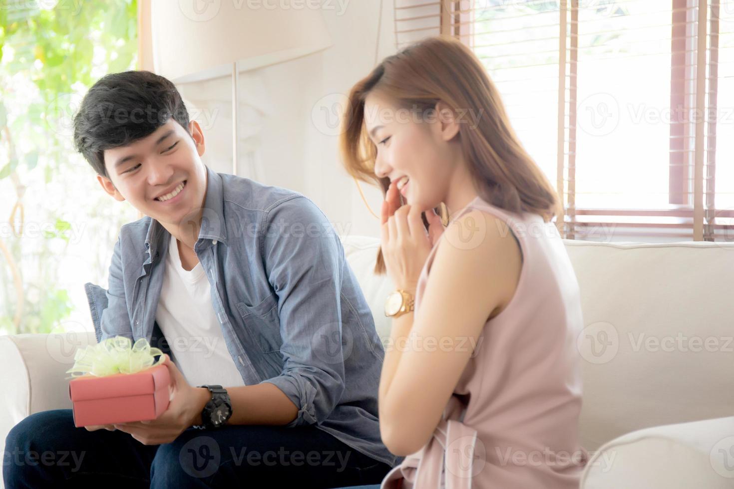 una joven pareja asiática celebra el cumpleaños juntos, un hombre asiático le da una caja de regalo a una mujer para sorprenderla en la sala de estar, una mujer se siente feliz y emocionada por el aniversario, el concepto de san valentín. foto