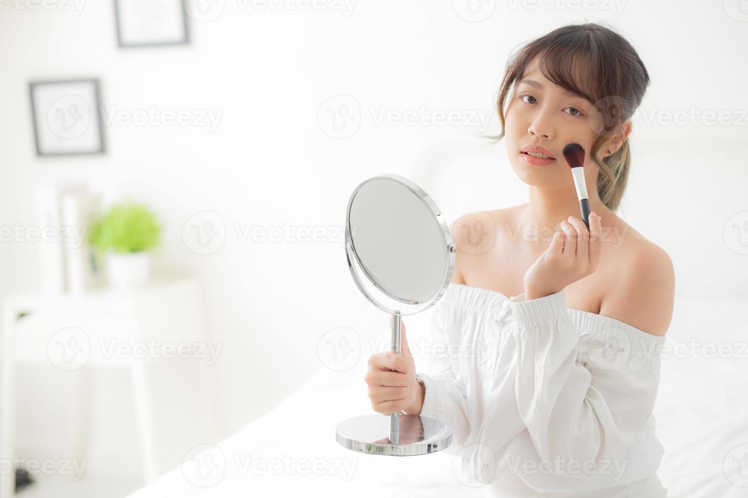 Beauty portrait young asian woman smile with face looking mirror applying makeup with brush cheek in the bedroom, beautiful of girl holding blusher, skin care and cosmetic concept. photo
