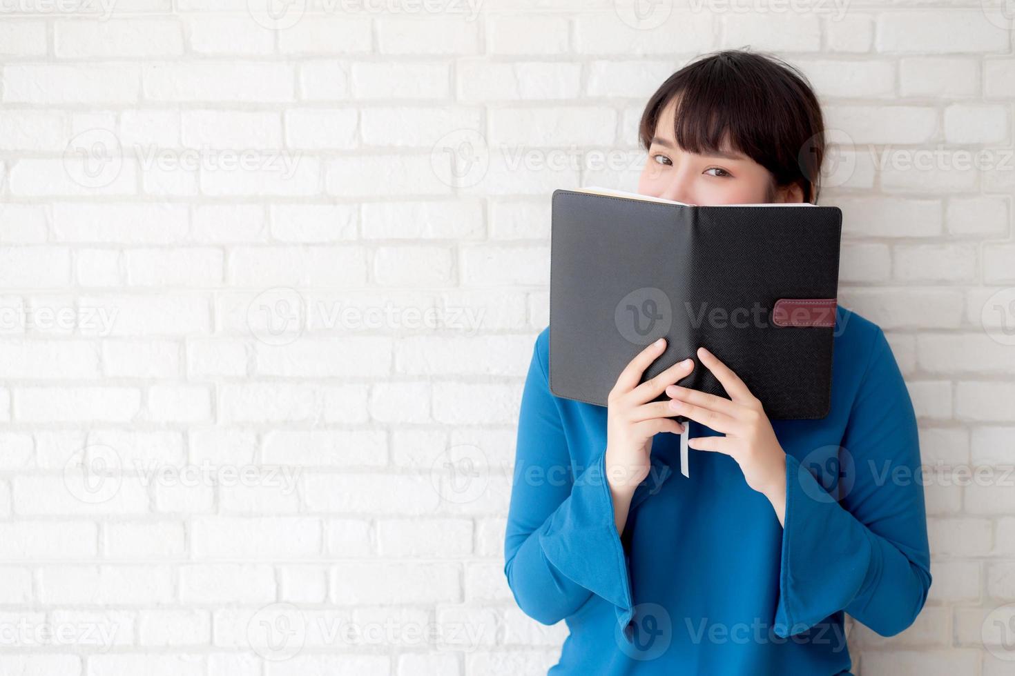 beautiful portrait young asian woman happy hiding behind covering the book with cement or brick concrete background, girl standing open notebook reading for learning, education and knowledge concept. photo
