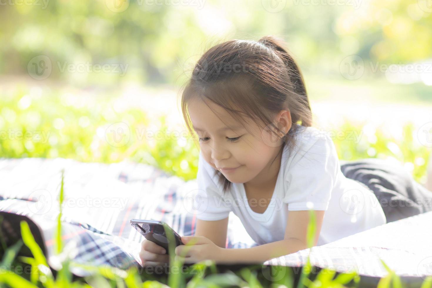 Beautiful asian child smiling using smart mobile phone in the garden, kid have passion playing game digital on smartphone on internet network in the park with happy, lifestyle and technology concept. photo