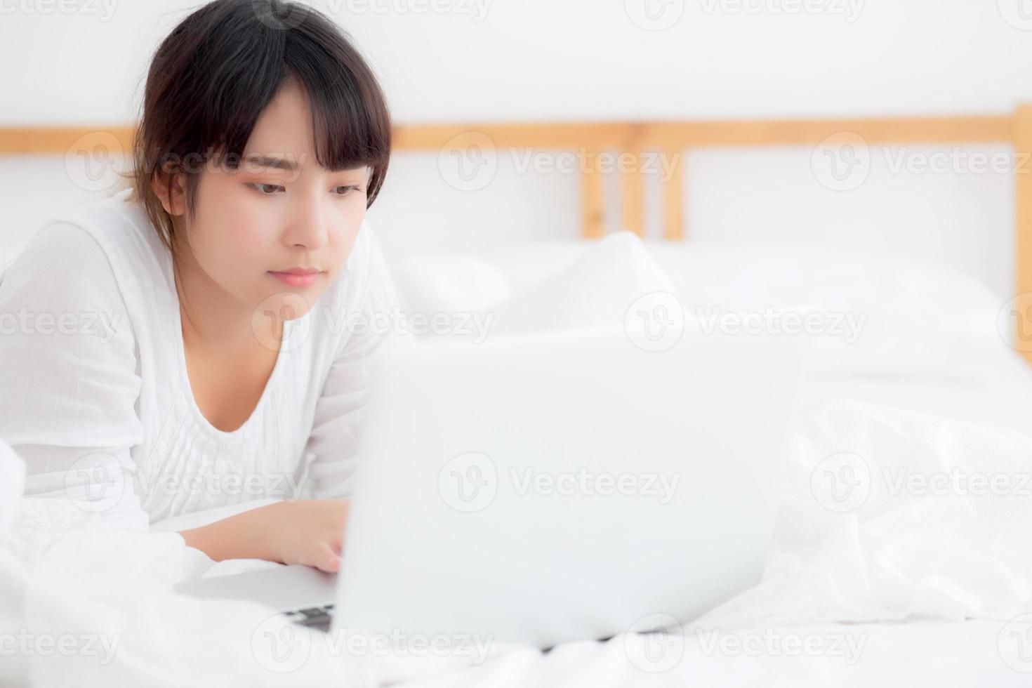 Beautiful portrait asian young woman smile lying on bed using laptop computer with happy at bedroom for leisure and relax, freelance with girl working notebook, communication concept. photo