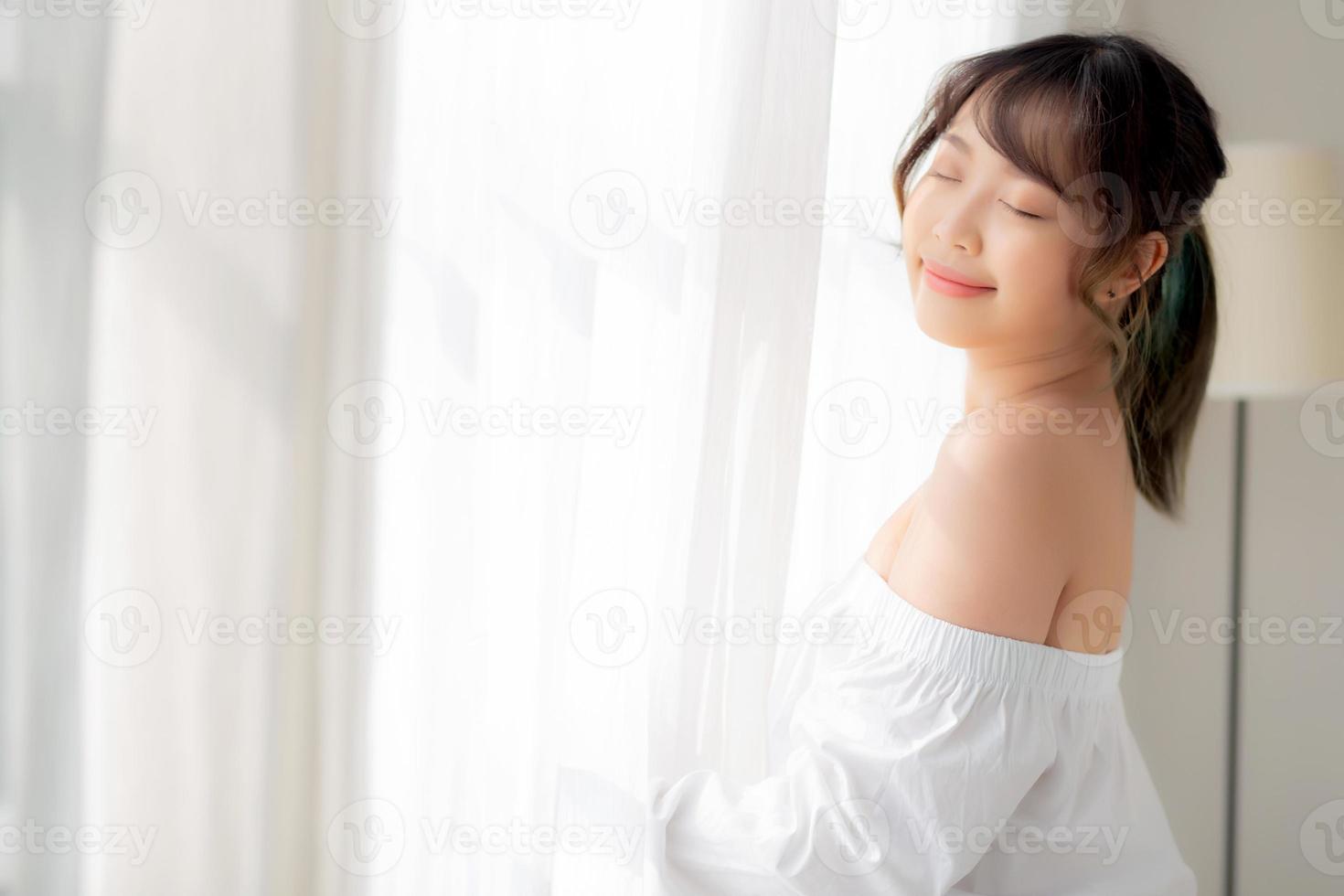 retrato de una hermosa joven asiática parada en la ventana y sonriendo mientras se despierta con el amanecer en la mañana, una chica feliz con frescura y alegre, estilo de vida y concepto de relajación. foto