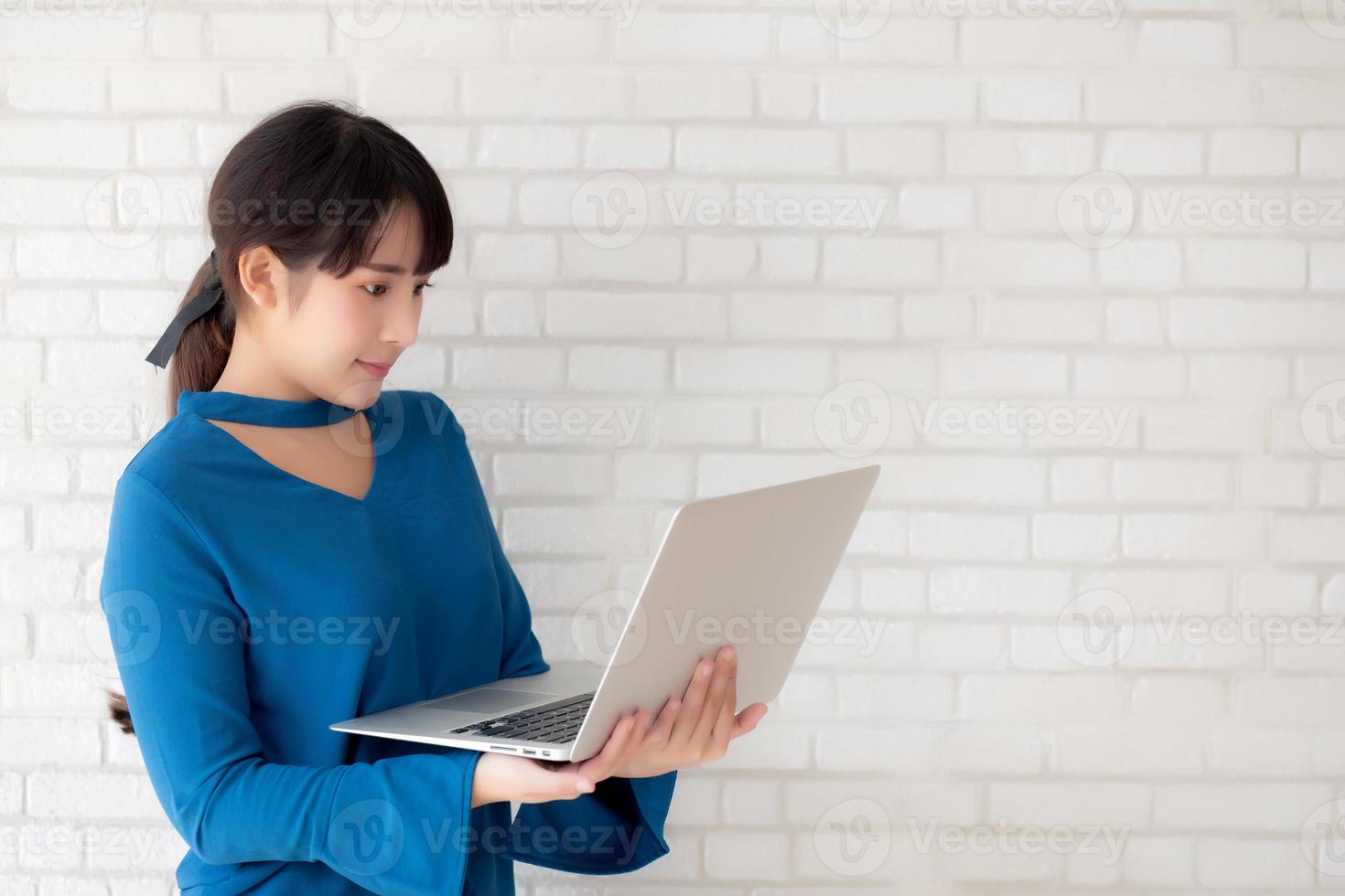 Beautiful portrait asian young woman smile using laptop standing at workplace on cement concrete background, girl happy with computer internet online, lifestyle and freelance business concept. photo