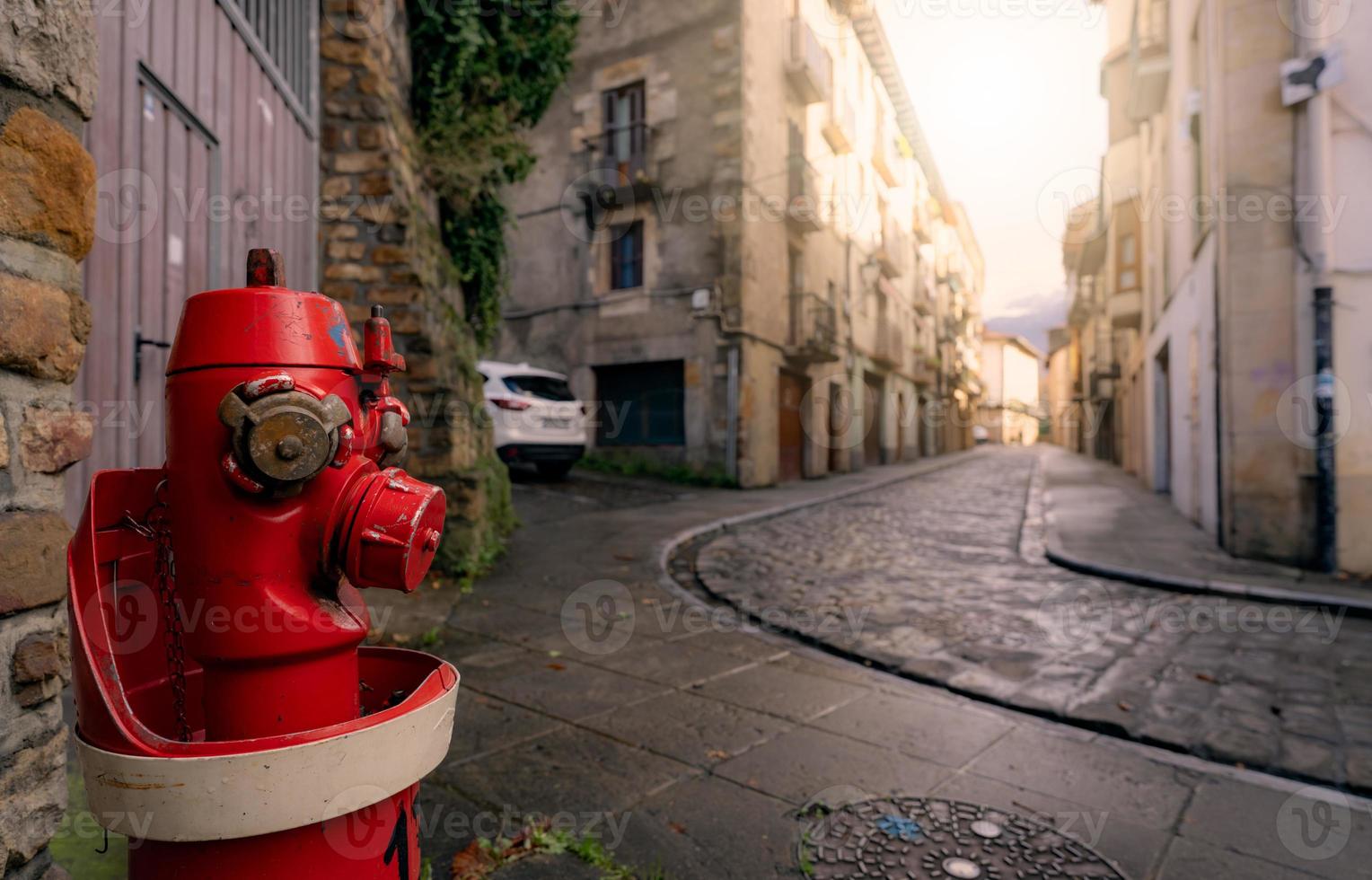 Red fire hydrant on sidewalk in Onati city, Spain. Fire hydrant on blur old building, white car, and street. Cityscape. Water supply for fire extinguisher. Fire control system of the city for safety. photo