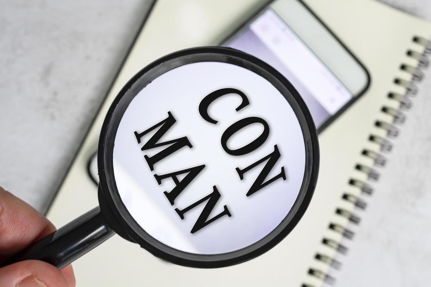 A man's hand holds a black magnifying glass by the handle with con man text. photo