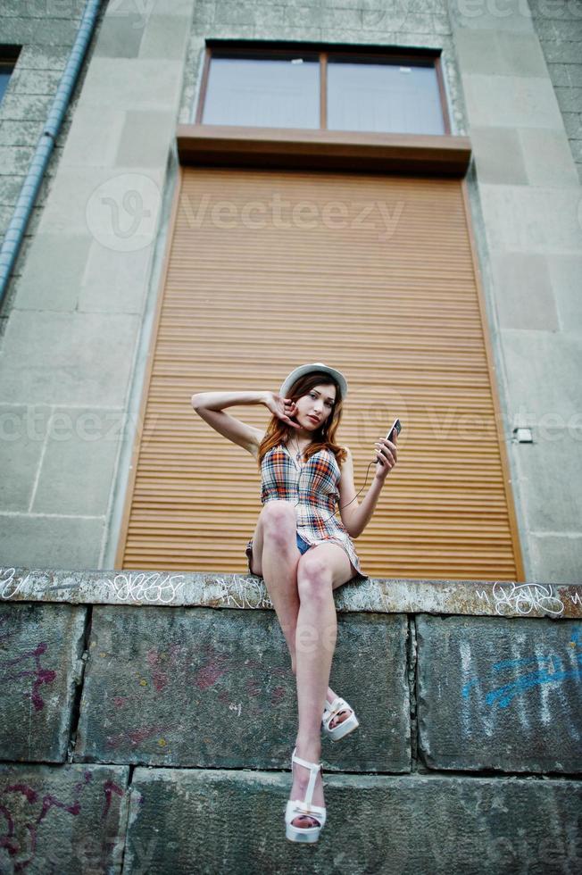 Amazing long legs with hig heels girl wear on hat against shutter listening music from headphones of mobile phone. photo