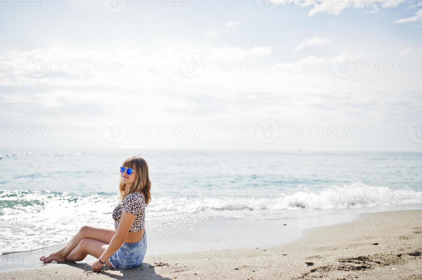 Beautiful model relaxing on a beach of sea, wearing on jeans short, leopard shirt and sunglasses. photo
