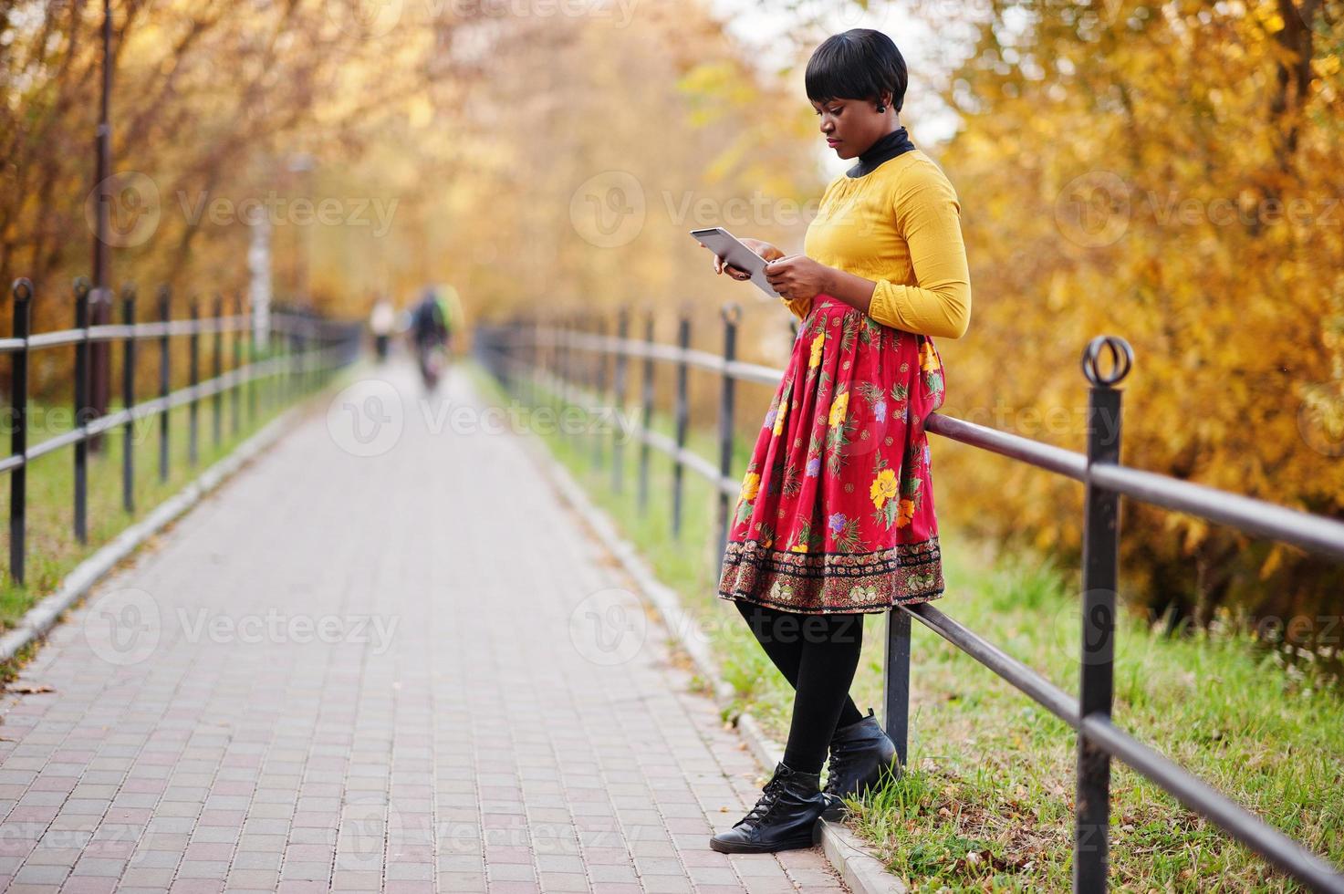 niña afroamericana con vestido amarillo y rojo en el parque de otoño dorado con tableta en las manos. foto