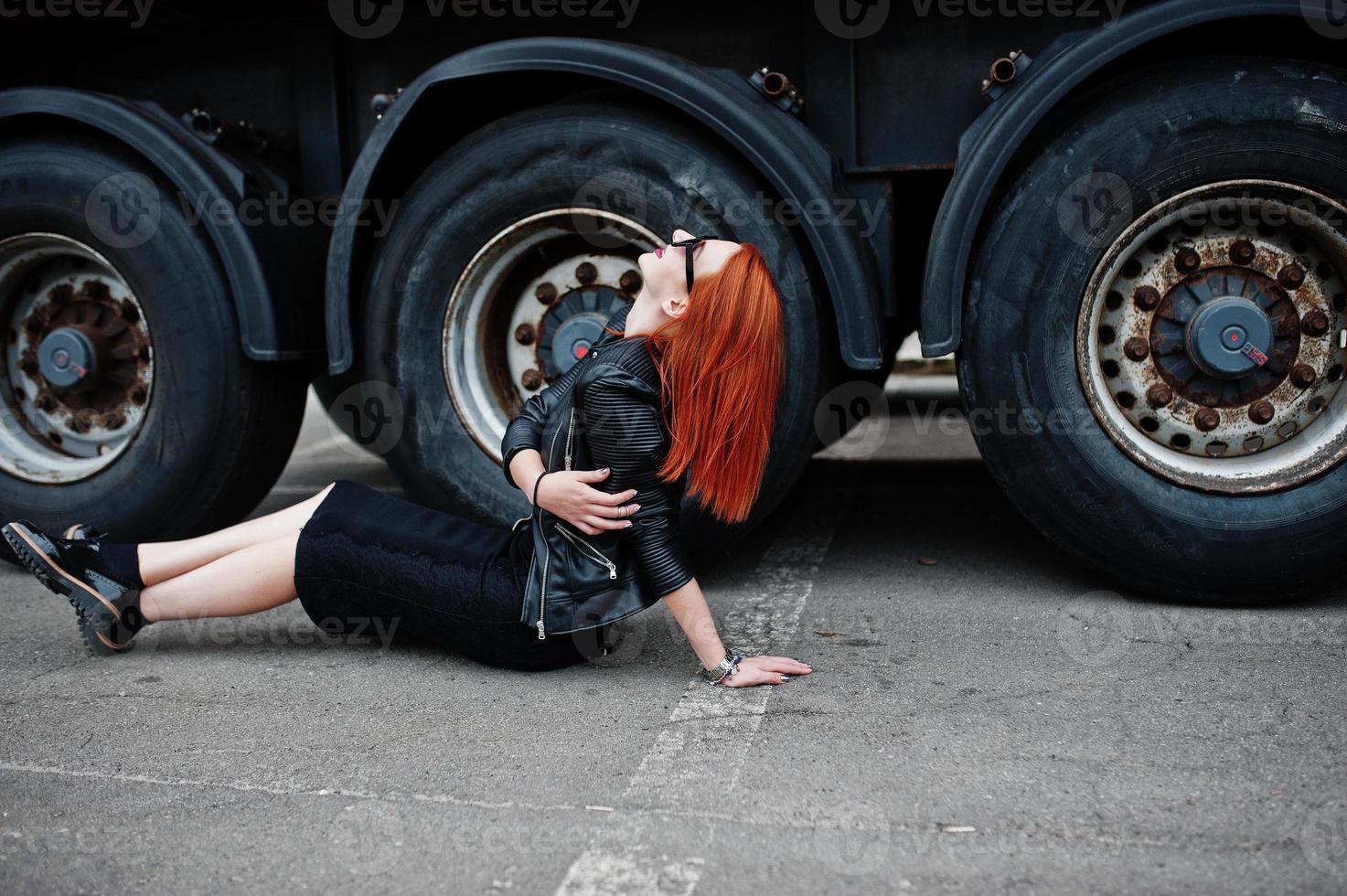 Red haired stylish girl wear in black, sitting against large truck wheels. photo