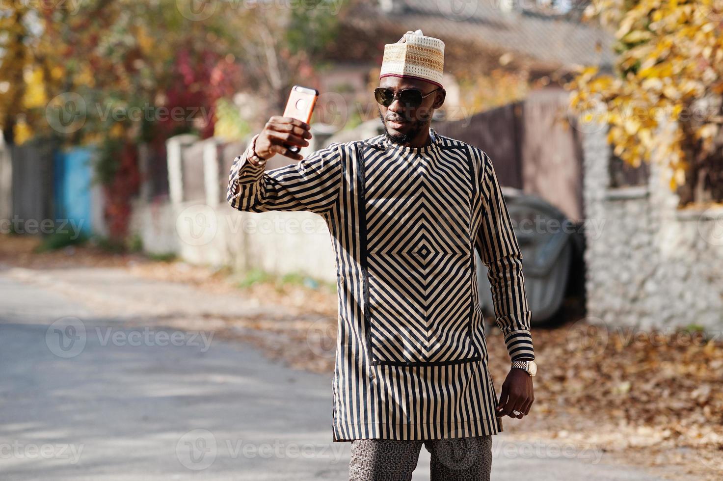 Portrait of stylish black african american man at hat and sunglasses against sunny autumn fall background doing selfie from mobile phone. Rich people in africa at traditional dress. photo