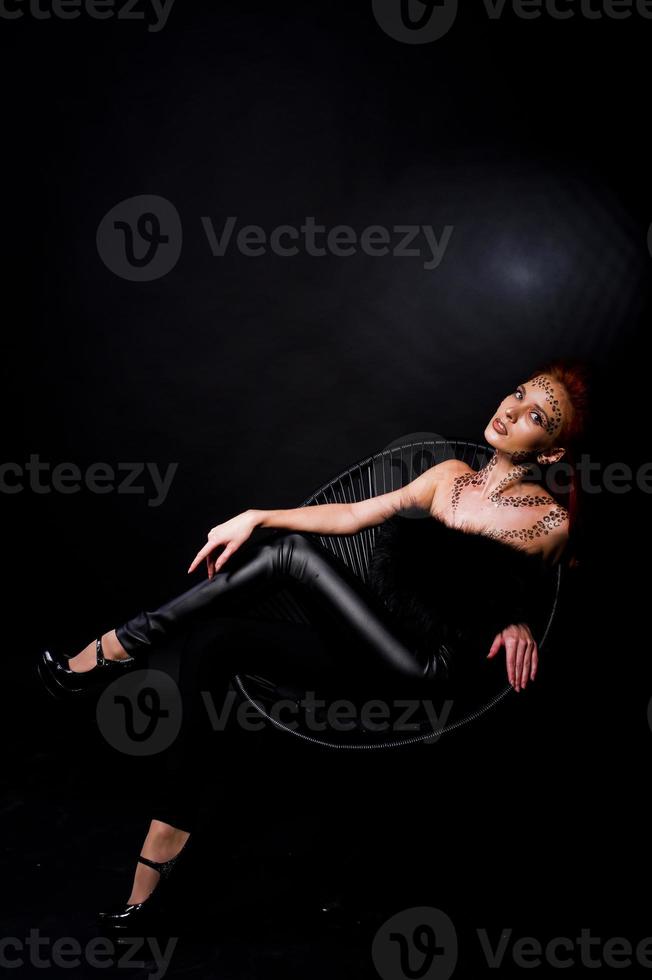 Fashion model red haired girl with originally make up like leopard predator isolated on black. Studio portrait on chair. photo