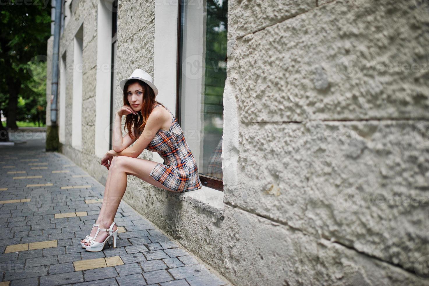 Amazing long legs with hig heels girl wear on hat sitting on the window sill on streets of city. photo